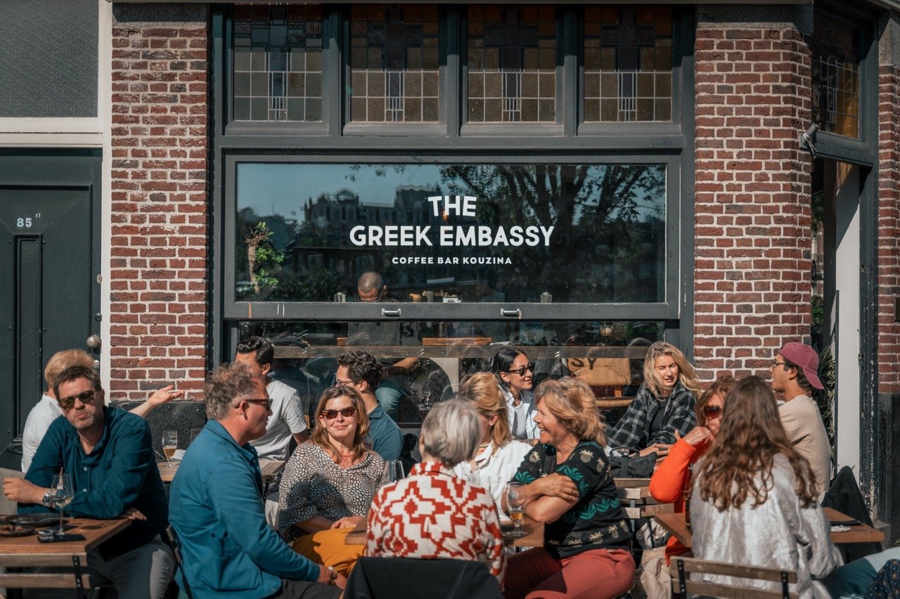 Buitenrestaurant voor de Greek Embassy, met groepen mensen die aan tafels zitten en genieten van de zon.