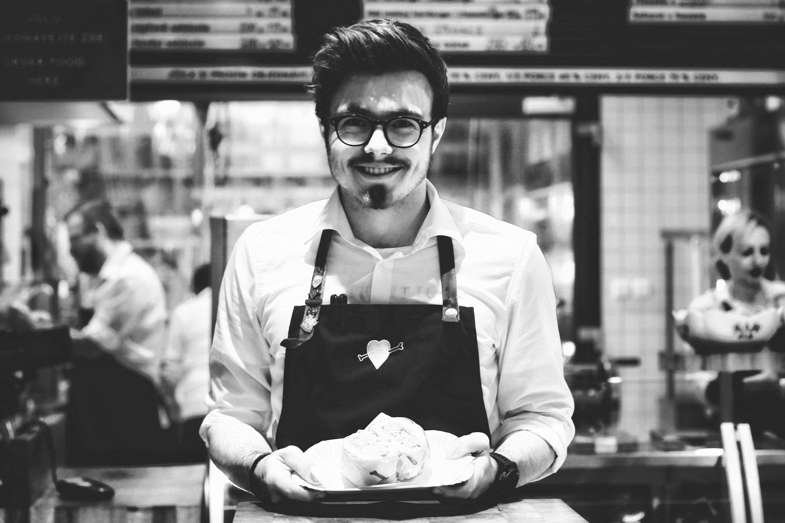 Man met bruin haar, bril, witte shirt en zwarte schort met hart en pijl in een restaurant, die een bord met eten vasthoudt.