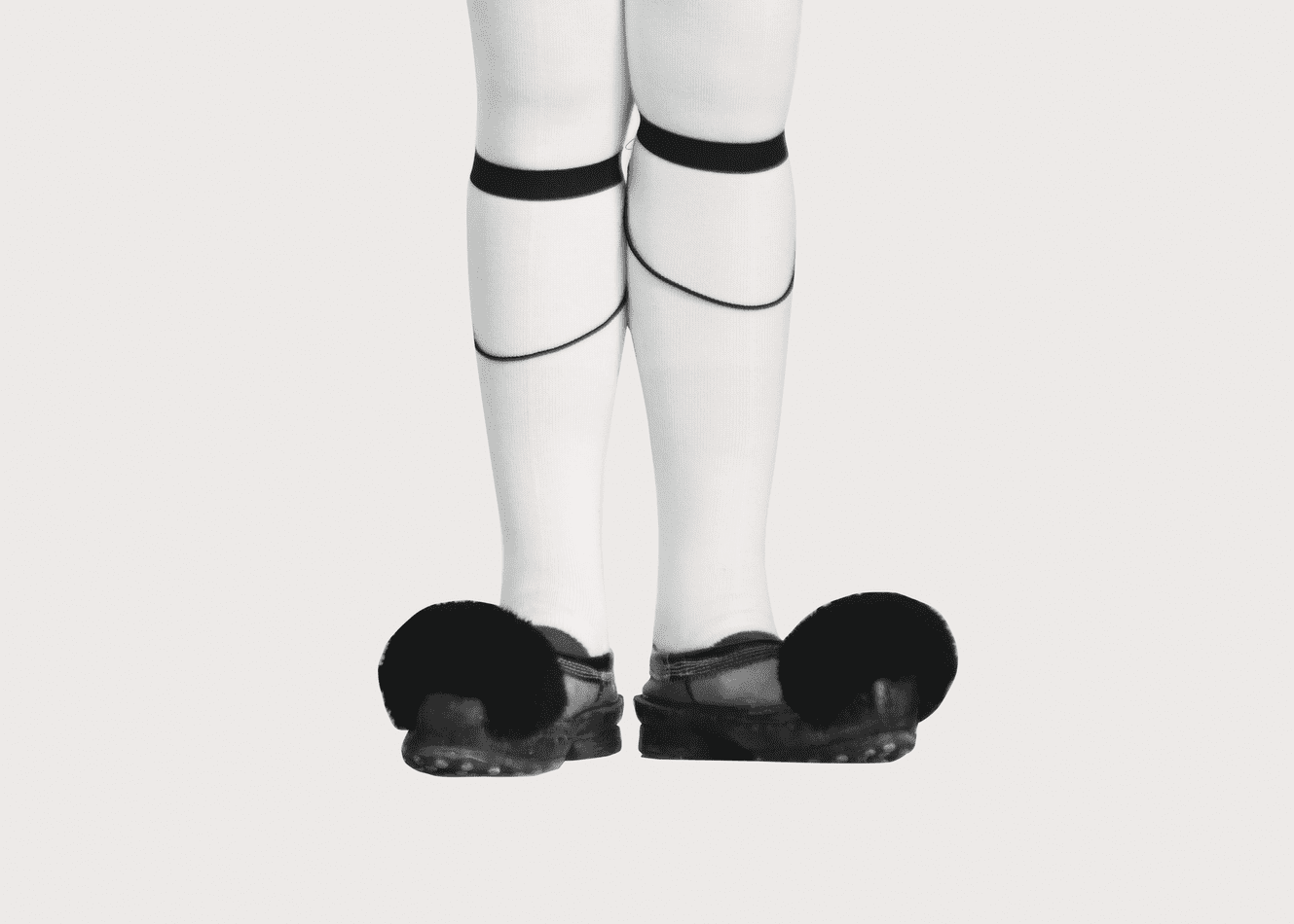 De onderkant van een persoon in witte sportkousen en rubberen slippers, met zwarte banden onder de knieën en rondom de benen.