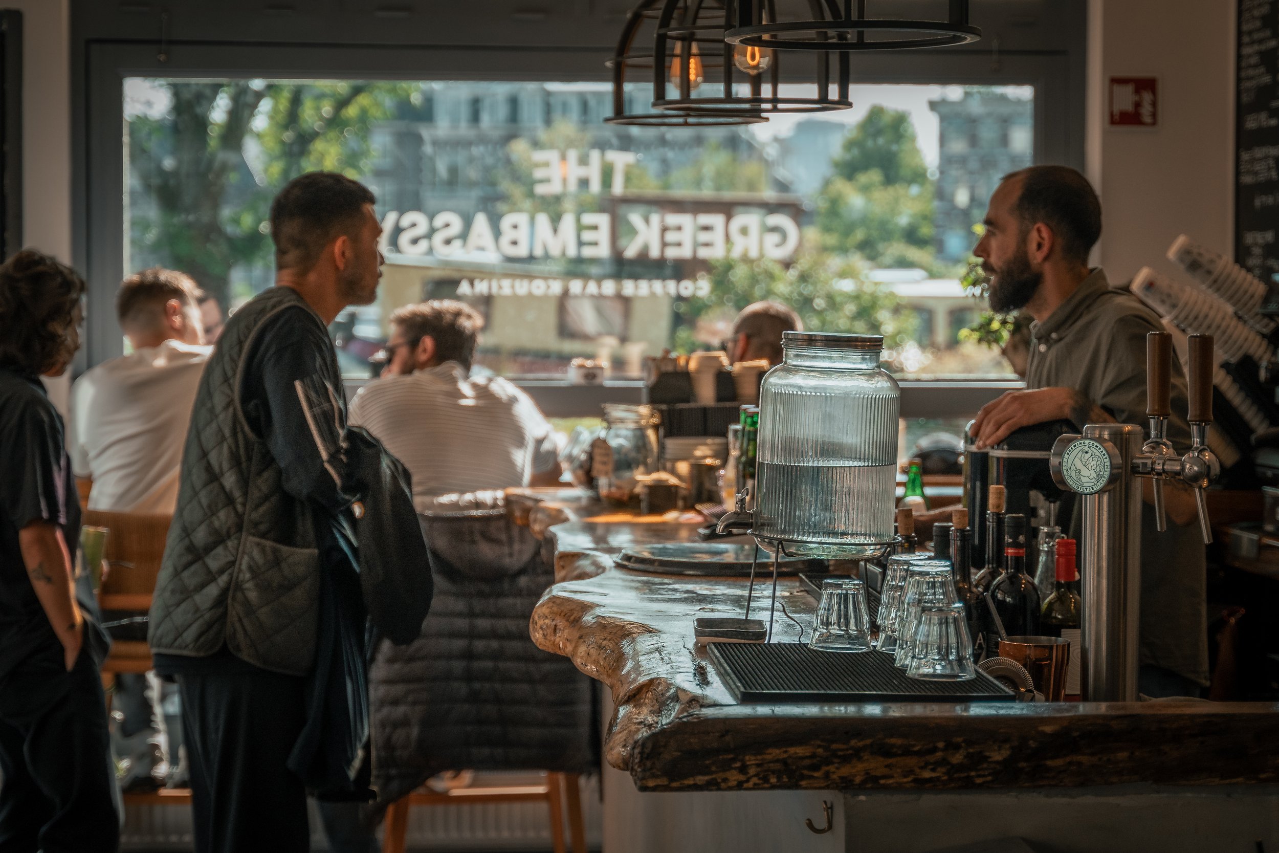 Binnen in een café met een houten bar, waarop glazen en een waterkan staan, en mensen die een drankje bestellen en genieten.