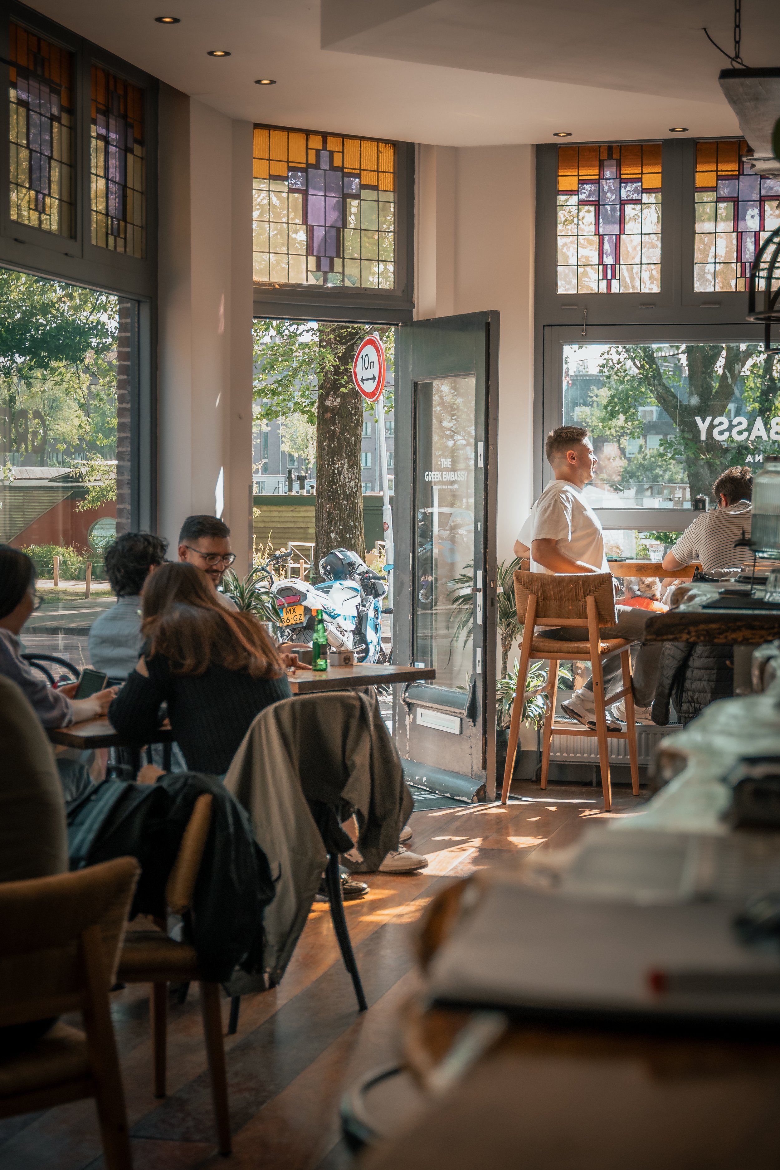 Binnen in een gezellige café met mensen die praten en genieten, grote ramen met uitzicht op de straat en kleurrijke glas-in-loodramen aan het plafond.
